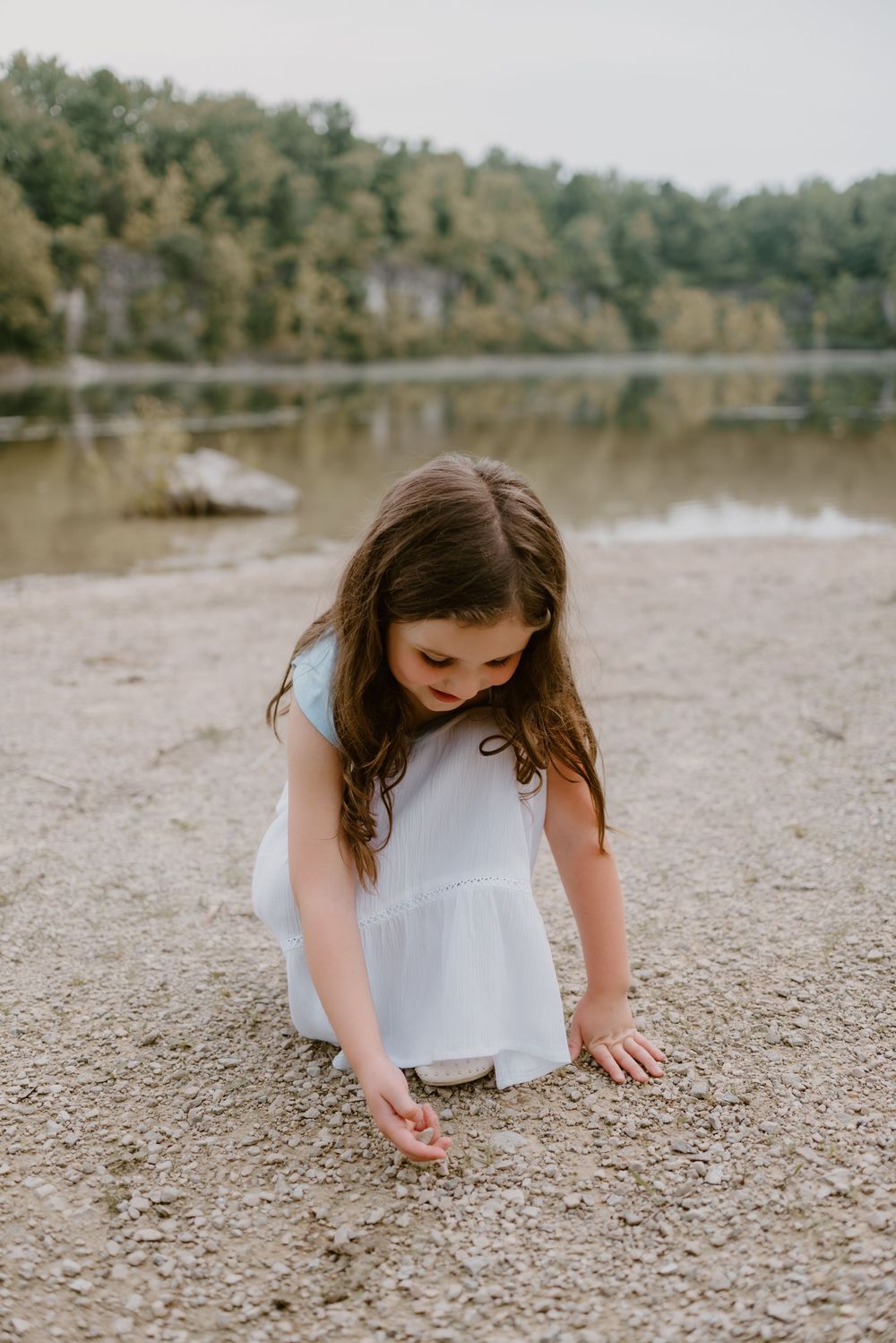 Young girl in white dress exploring rocky shore by lake.