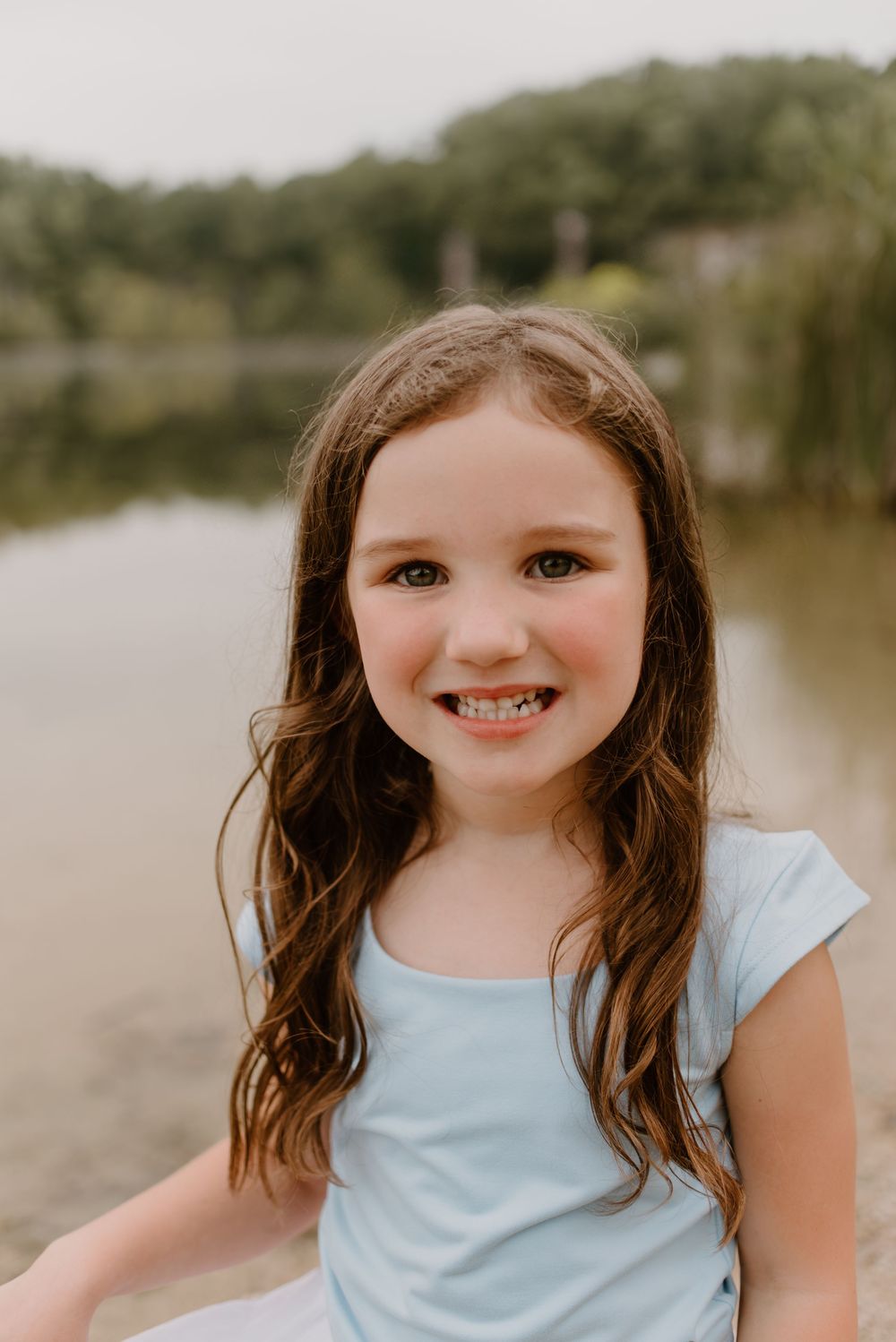 A young person in a light blue shirt smiles warmly against a lakeside backdrop.