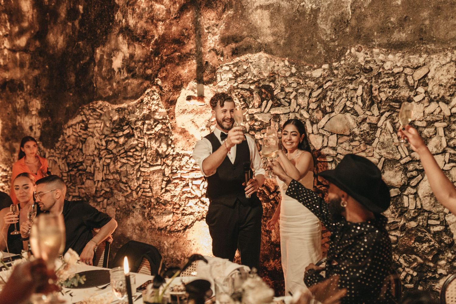Wedding reception scene in rustic cave setting with guests raising glasses.