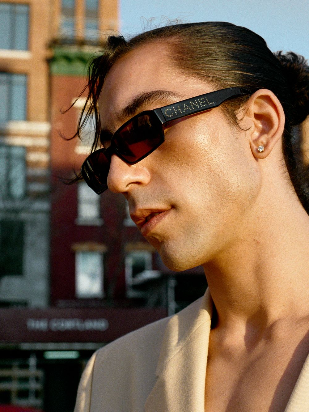 Person in Chanel sunglasses and beige blazer poses against urban backdrop with red brick building in soft sunlight.