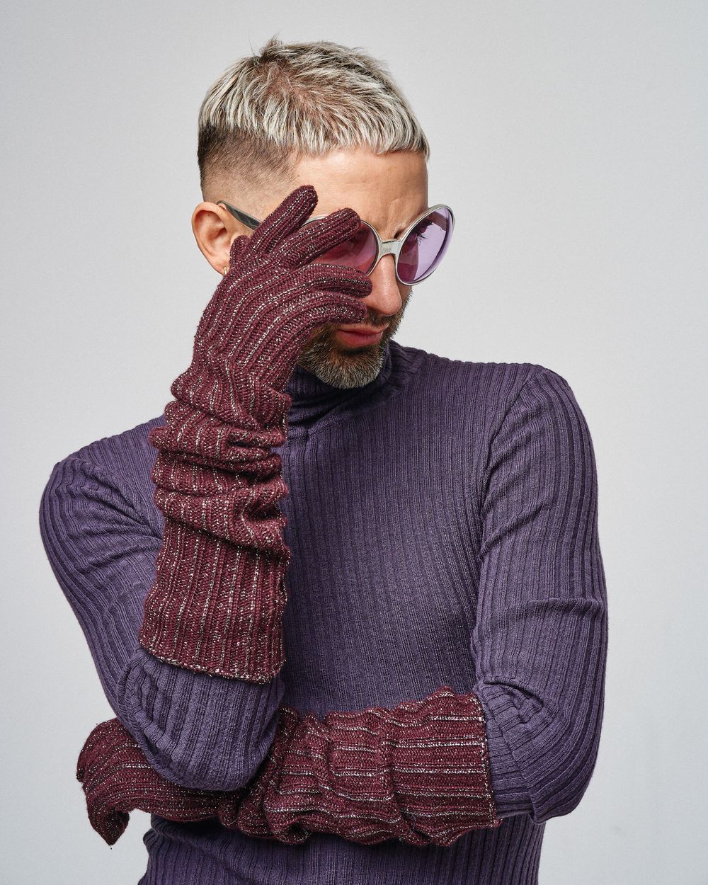 Person in purple sweater wearing burgundy gloves and round sunglasses poses against grey background with hand near face.