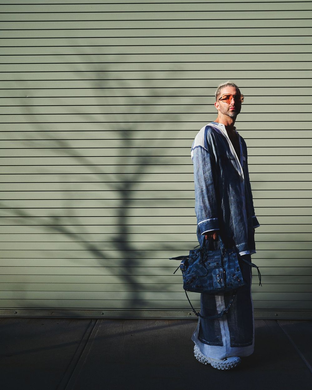 Person in denim jacket stands against green siding wall casting dramatic shadow in sunlight.