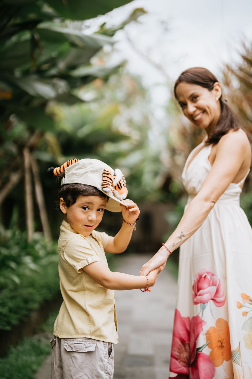 A young child in a hat holds hands with someone in a floral dress in a tropical garden setting.