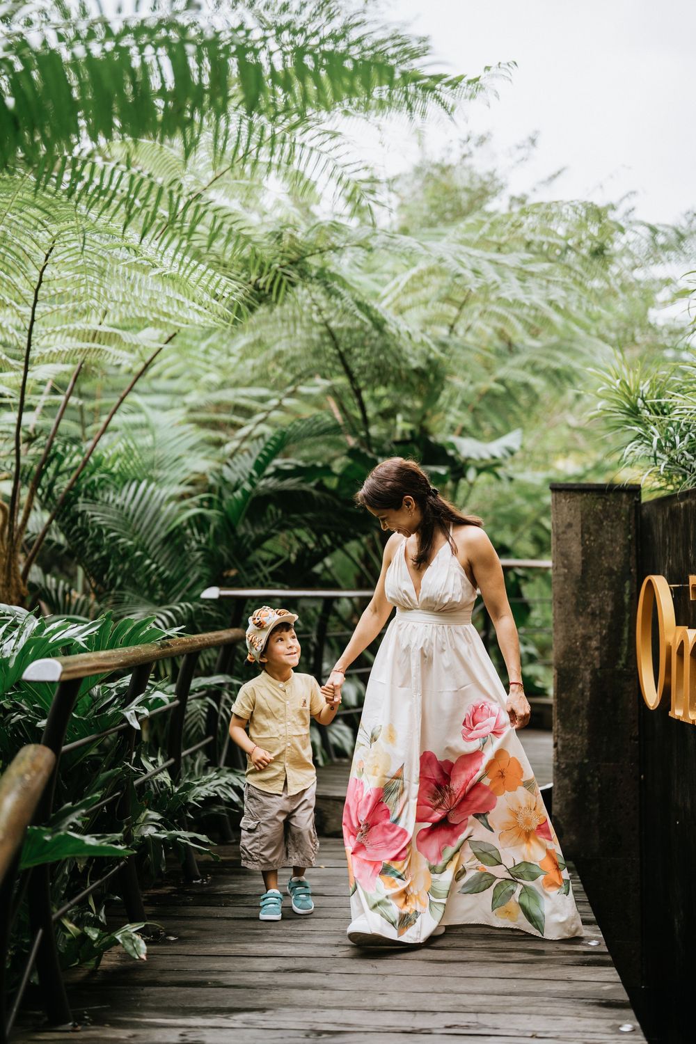 A person in a floral maxi dress walks with a child on a wooden pathway surrounded by tropical plants.