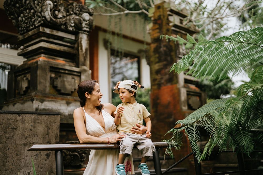 A pair share a tender moment in a lush tropical garden setting with traditional Balinese architecture.