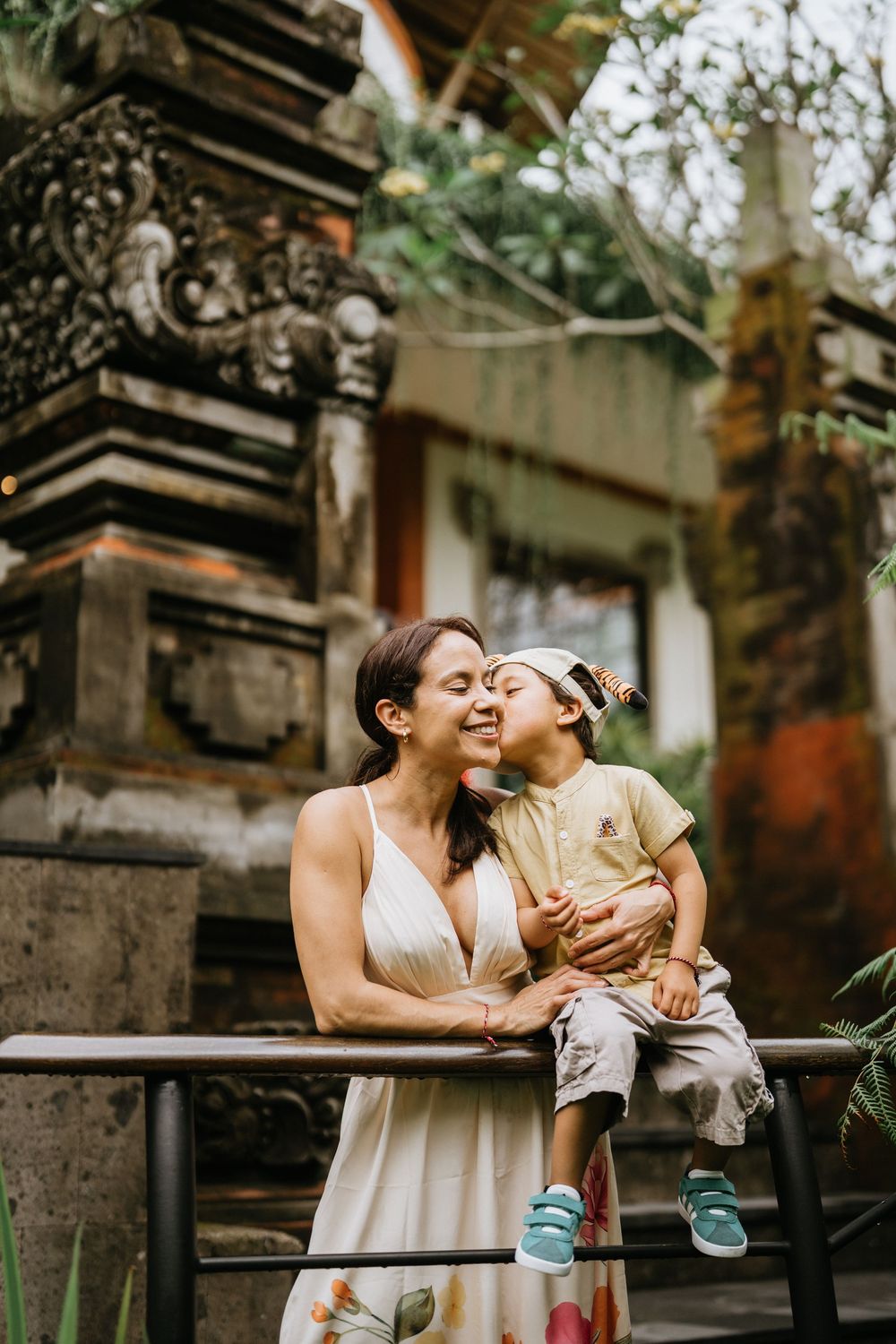 Two figures embrace while sitting on stone steps surrounded by tropical foliage and traditional architecture.