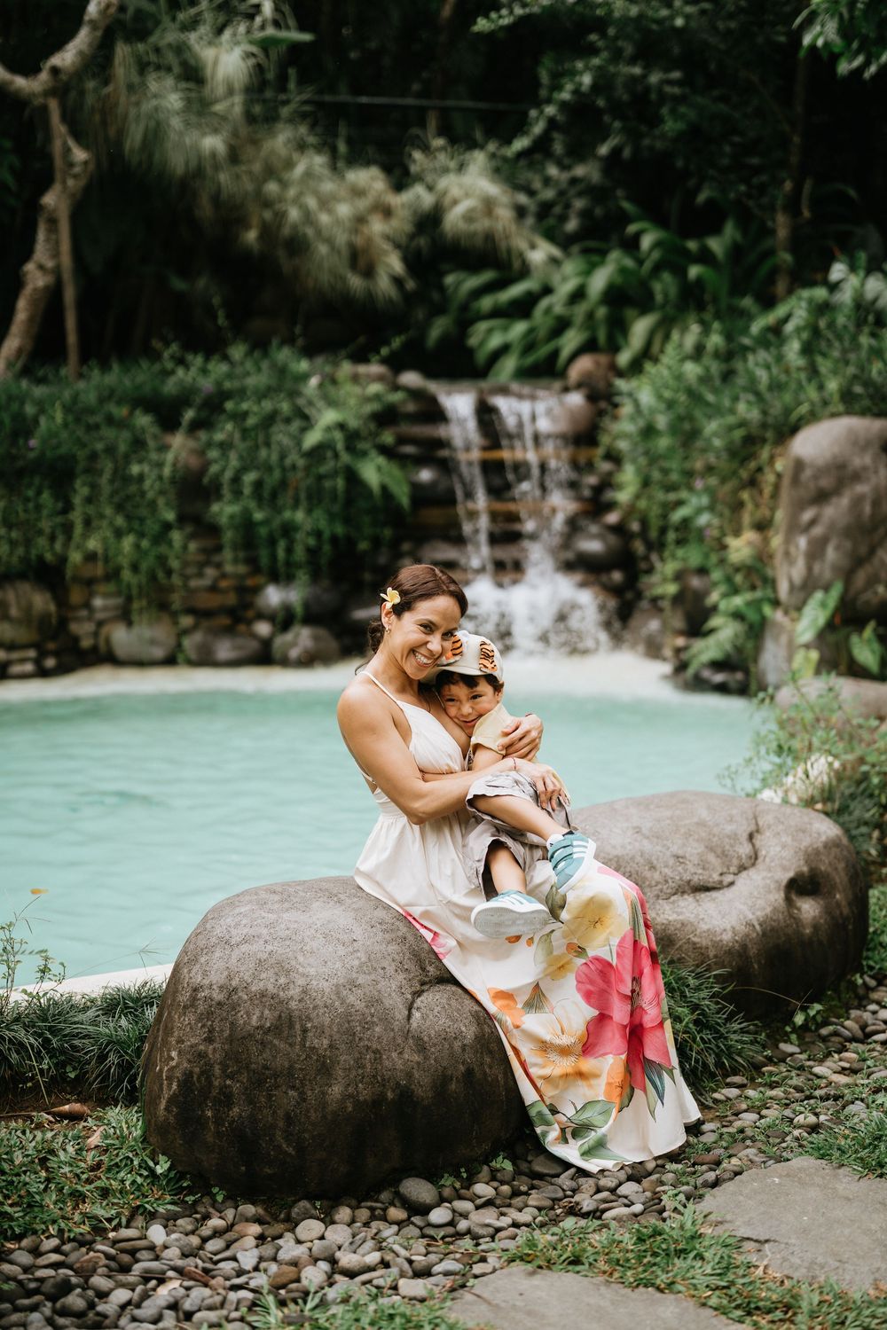 A pair relaxes on large rocks beside a turquoise pool surrounded by lush tropical vegetation.