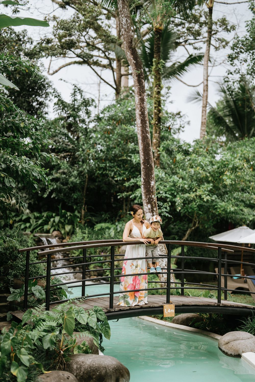 A wooden bridge curves over turquoise water in a lush tropical garden setting.