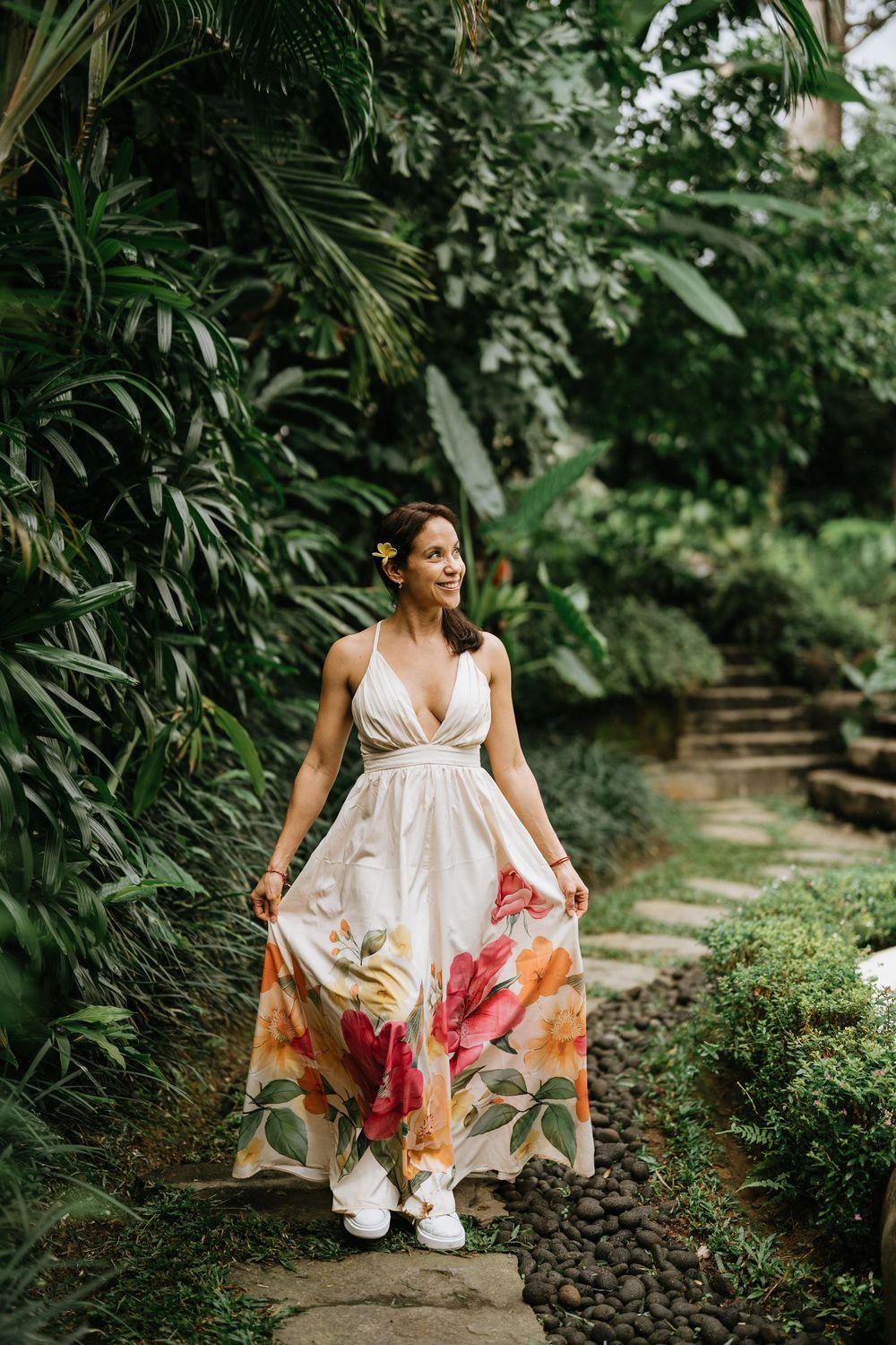 A person in a white floral dress poses in a tropical garden setting with stone pathways.