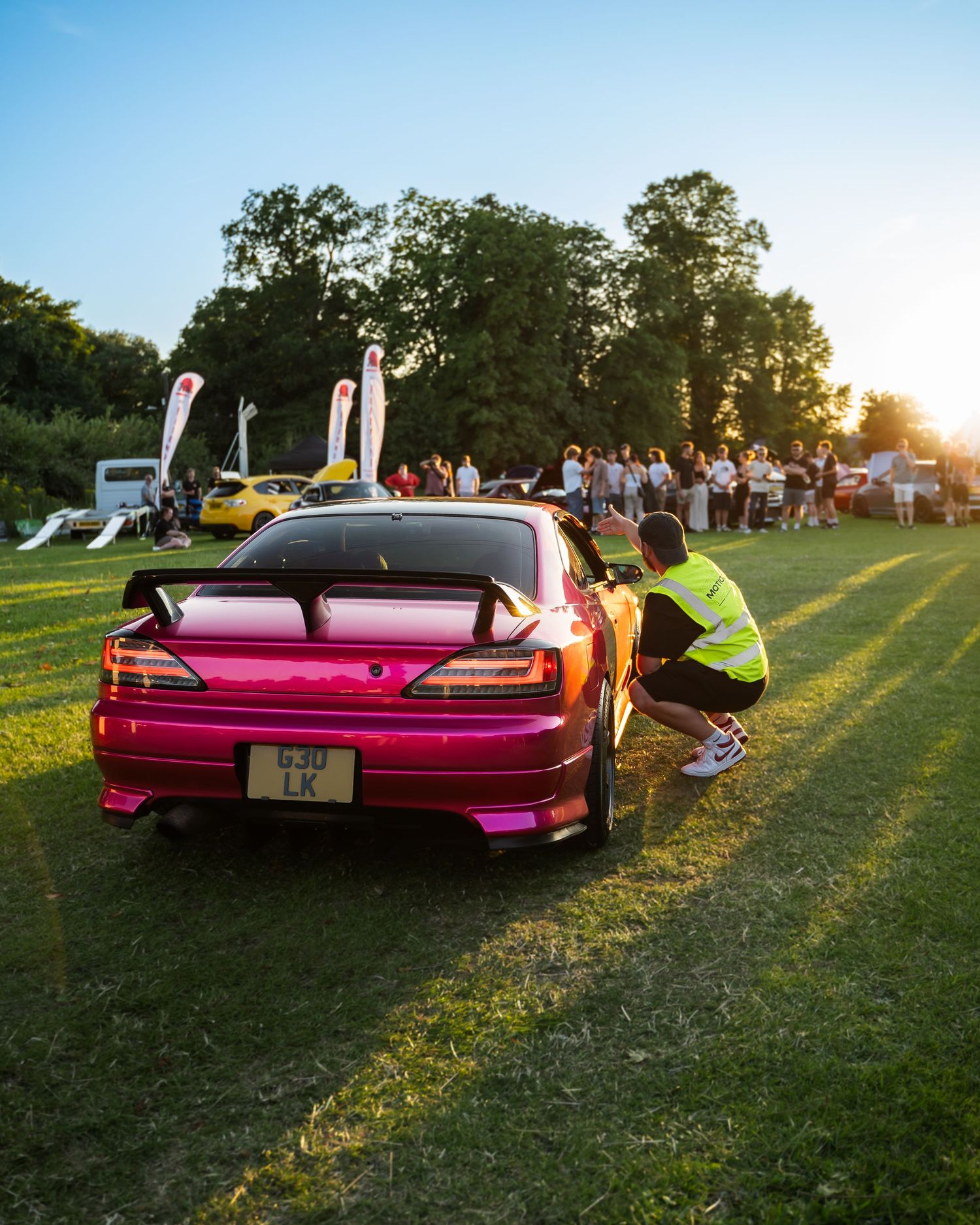 Pink vintage car displayed at an outdoor event during golden hour sunset.