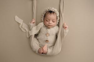 Sleeping newborn baby in white onesie rests peacefully in beige fabric sling during professional photography session.