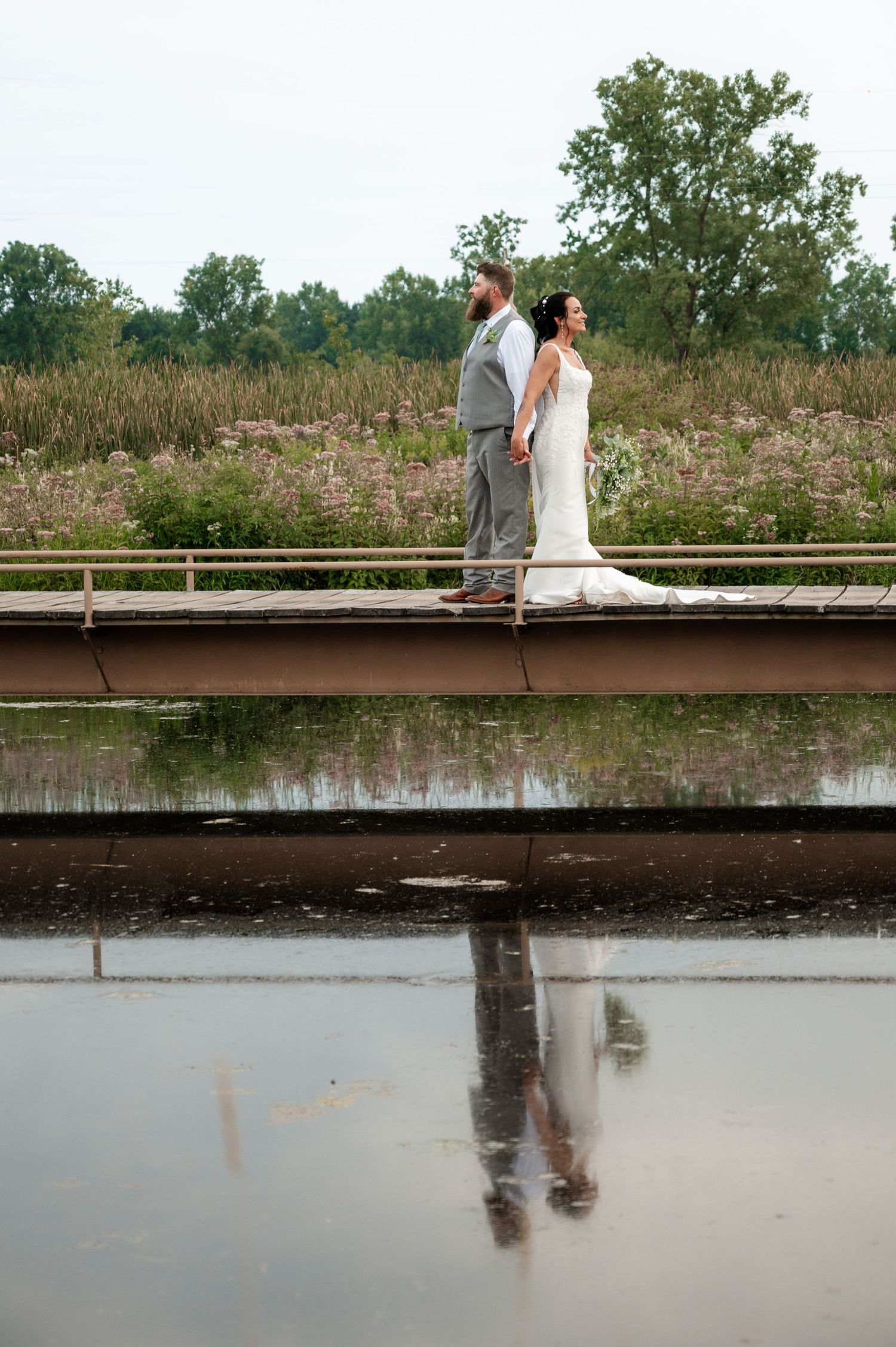 Bride and groom standing back-to-back on bridge reflected in water with wildflower meadow.