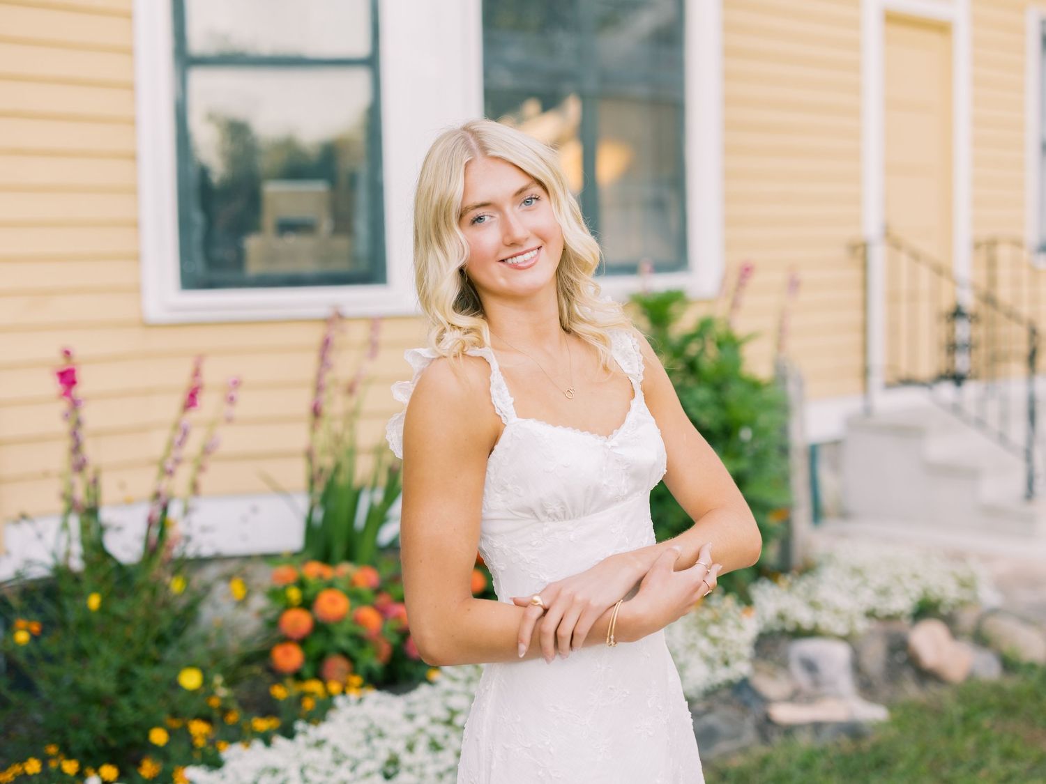 Person in white sundress standing outside yellow house with colorful garden flowers in summer setting.