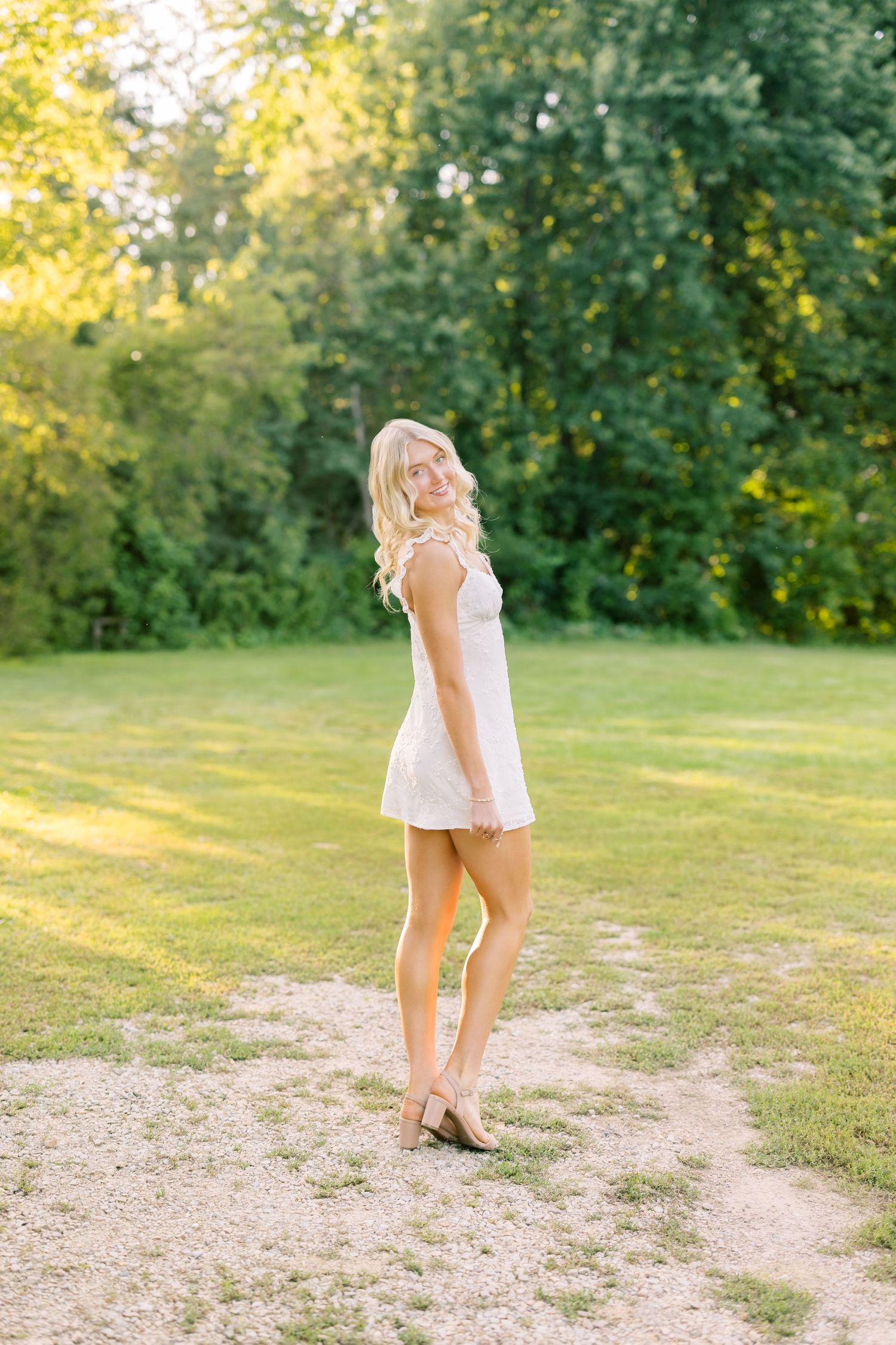 Summer sunset portrait session outdoors in a white dress and flowing hair against green trees and golden light.