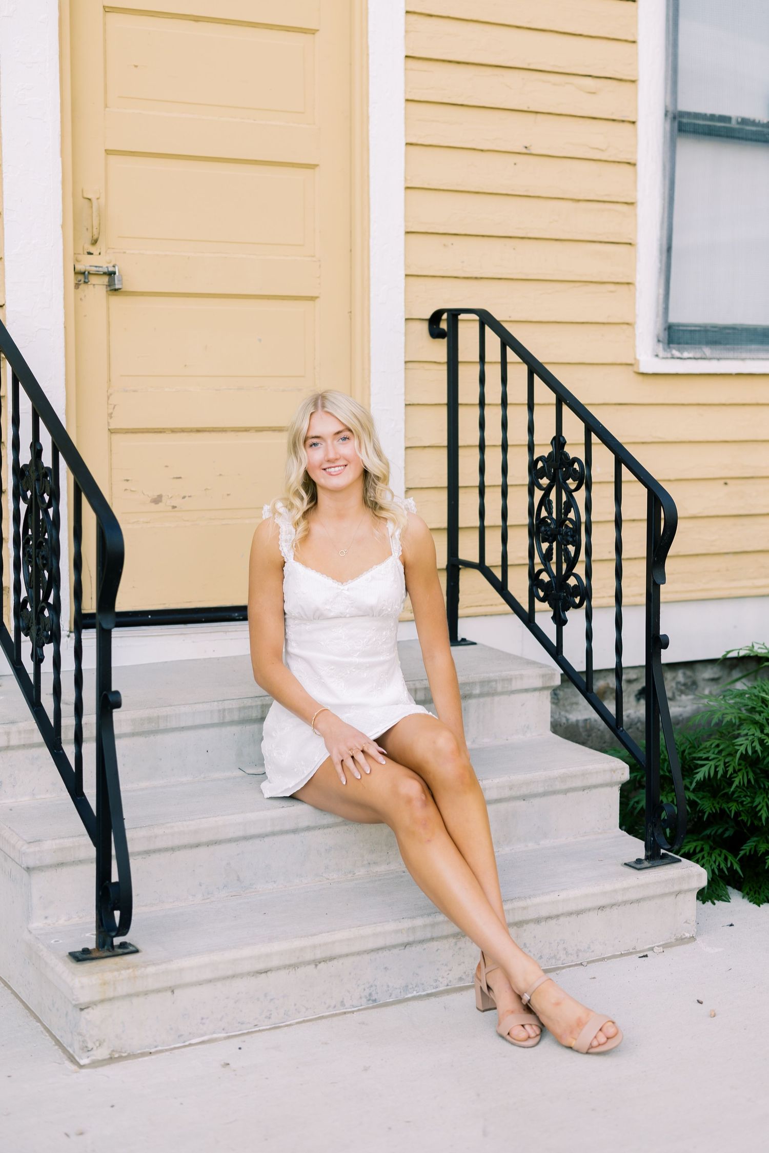 Person in white dress sits casually on concrete steps of yellow house with decorative black railings.