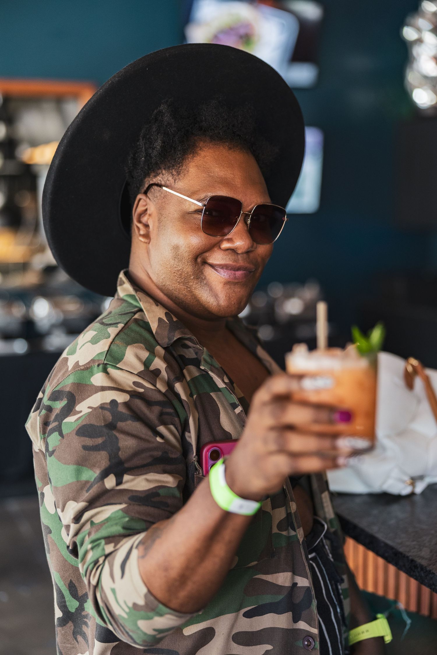 Person wearing camouflage jacket and sunglasses enjoys drinks at outdoor cafe setting.