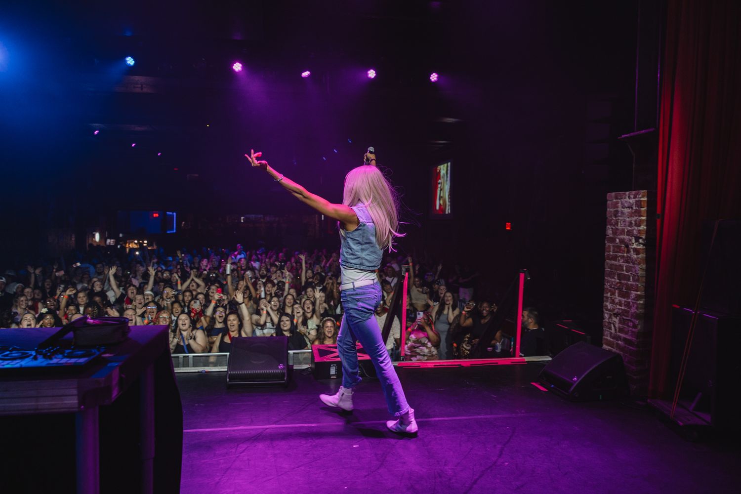 Blonde performer on stage with arms raised facing a large crowd in a purple-lit concert venue.