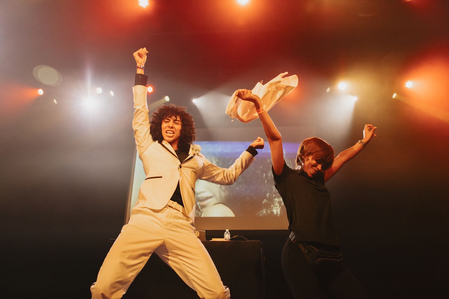 Two energetic performers celebrate on stage under colorful lights during a live concert performance.