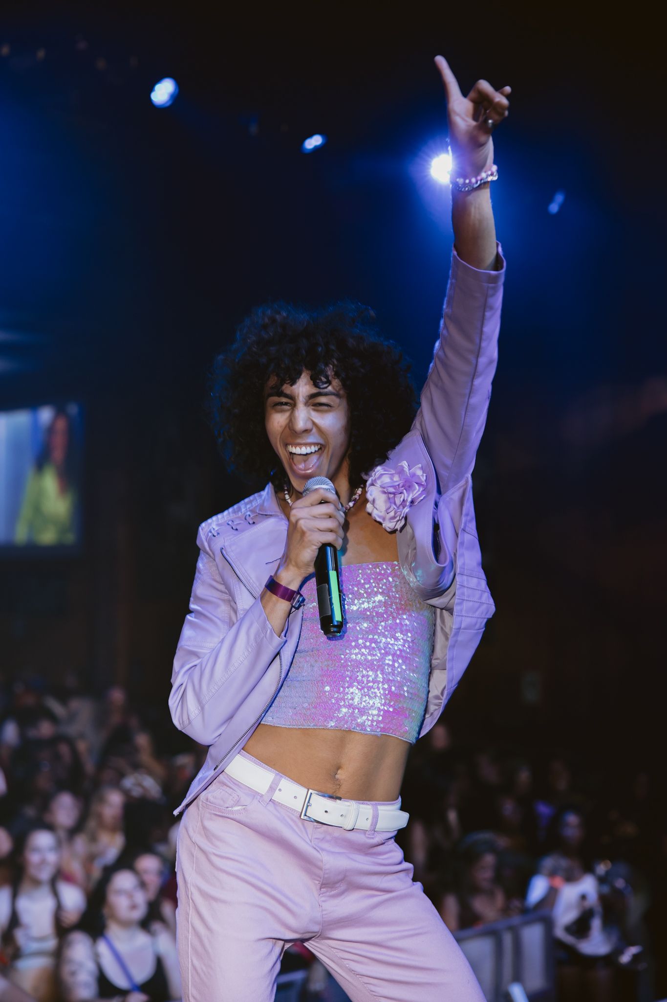 Energetic performer in pink sequin crop top and suit sings into mic on stage with arm raised.