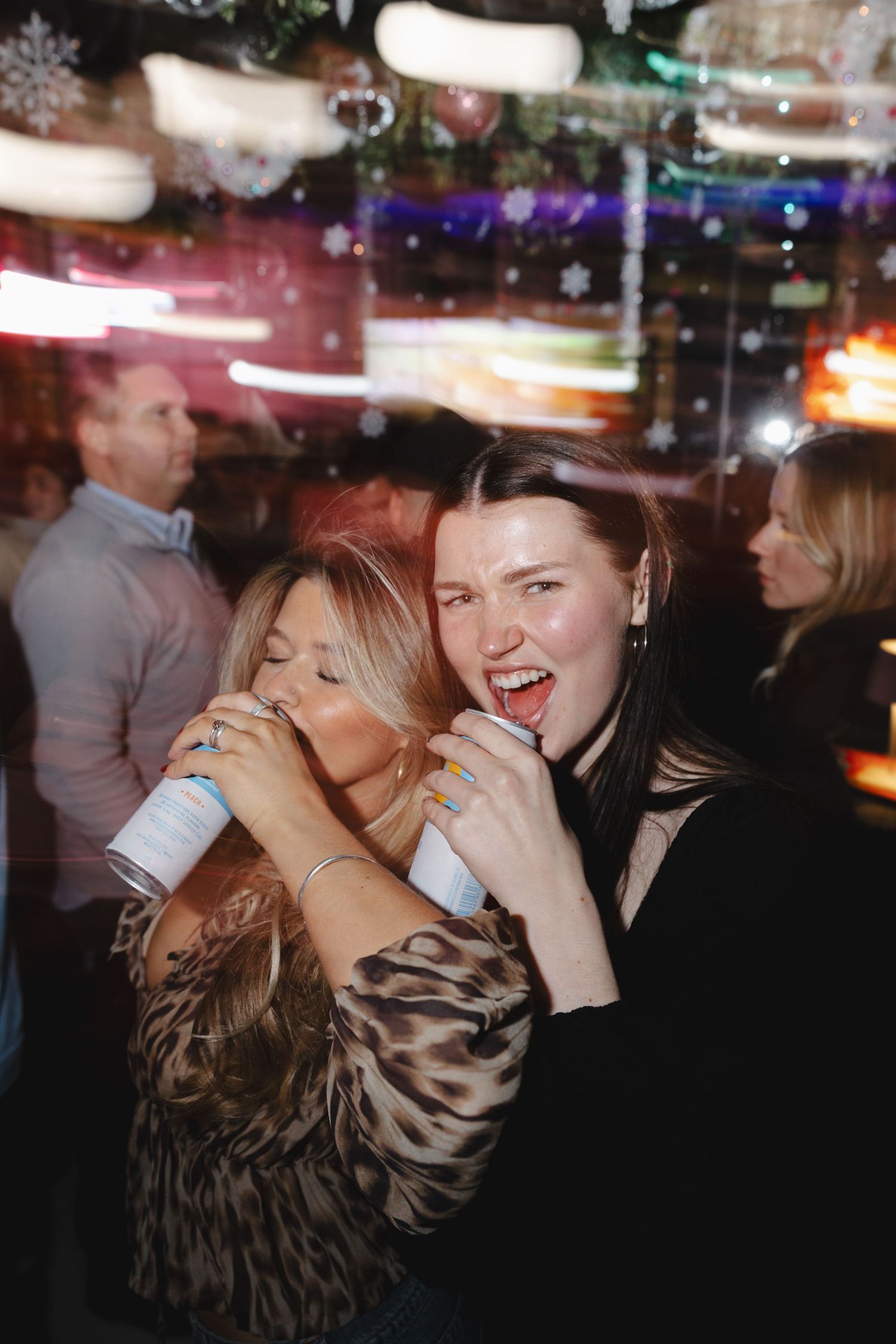 Two friends enjoying drinks and laughing together at a dimly lit nightclub with colorful lights in the background.