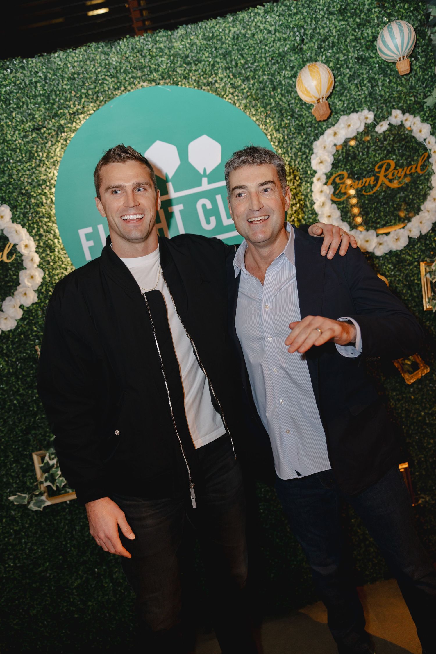 Two men in dark suits pose together at an event with a green hedge wall and decorative lighting in the background.