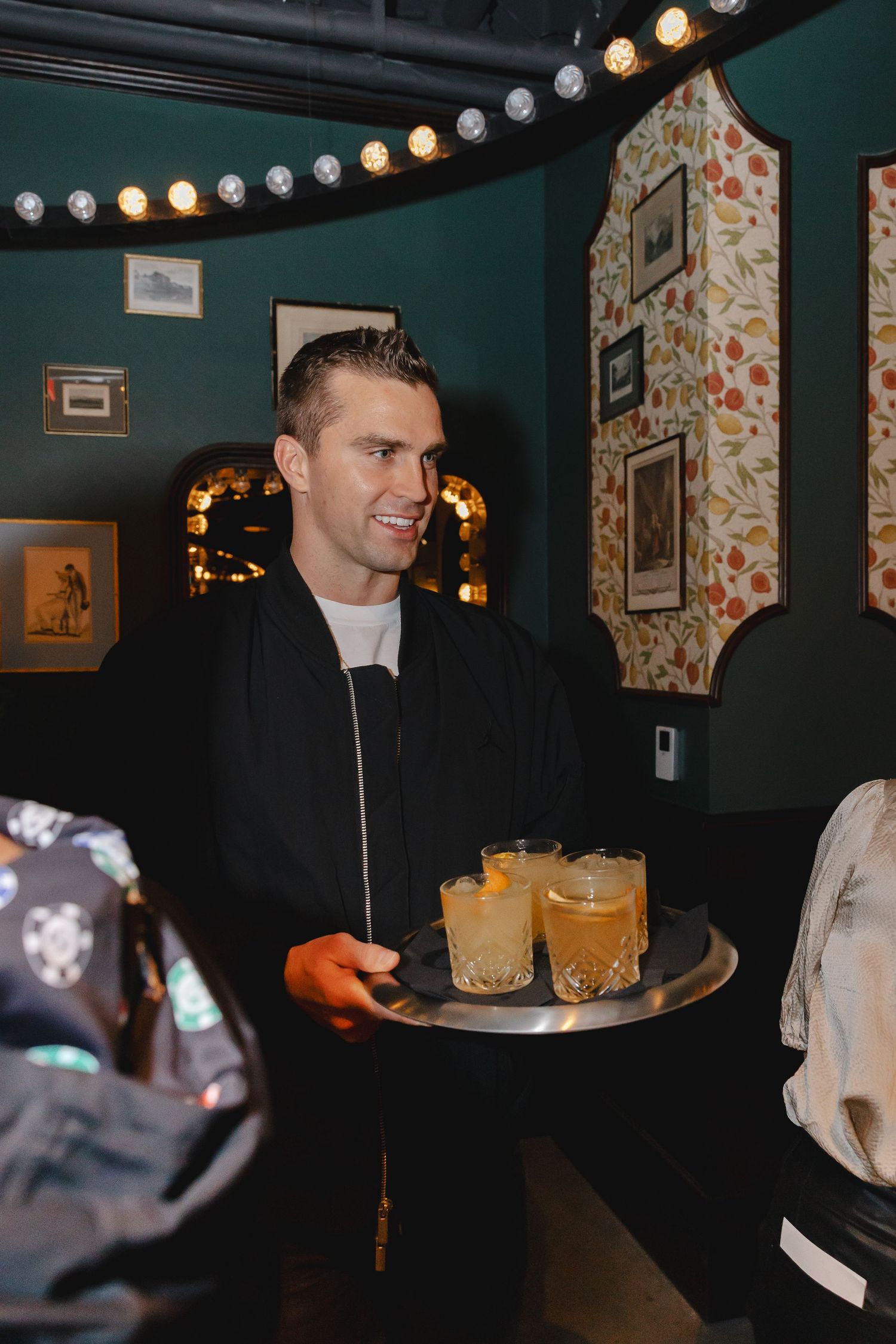 A dimly lit bar interior with string lights overhead and multiple orange cocktails being served.