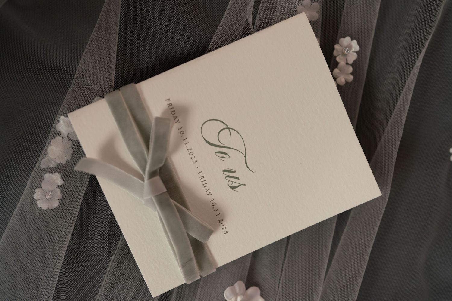 Elegant wedding invitation with script font, tied with a satin ribbon bow on sheer floral fabric.