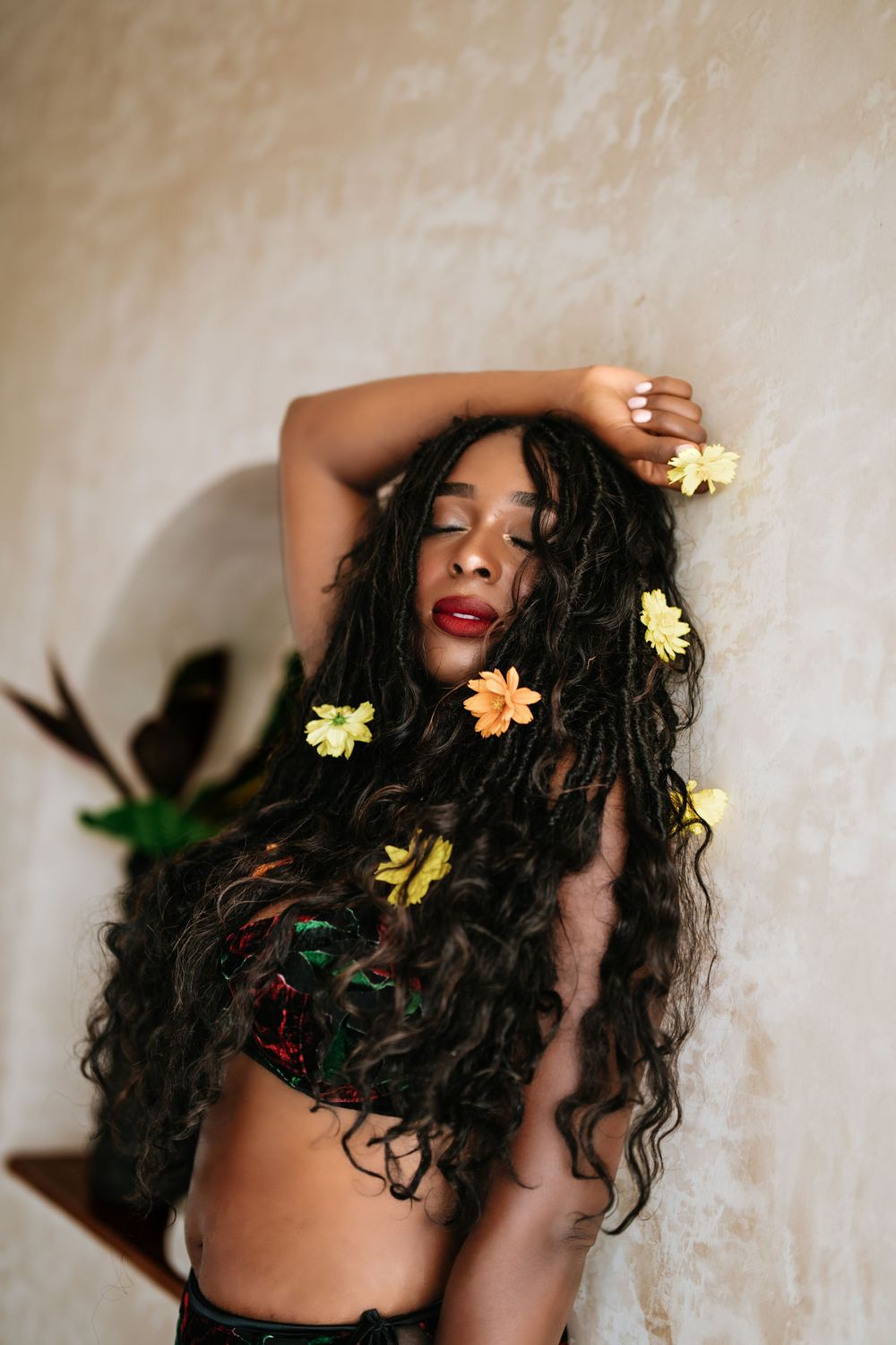 Artistic portrait with yellow flowers adorning long curly hair against neutral wall.