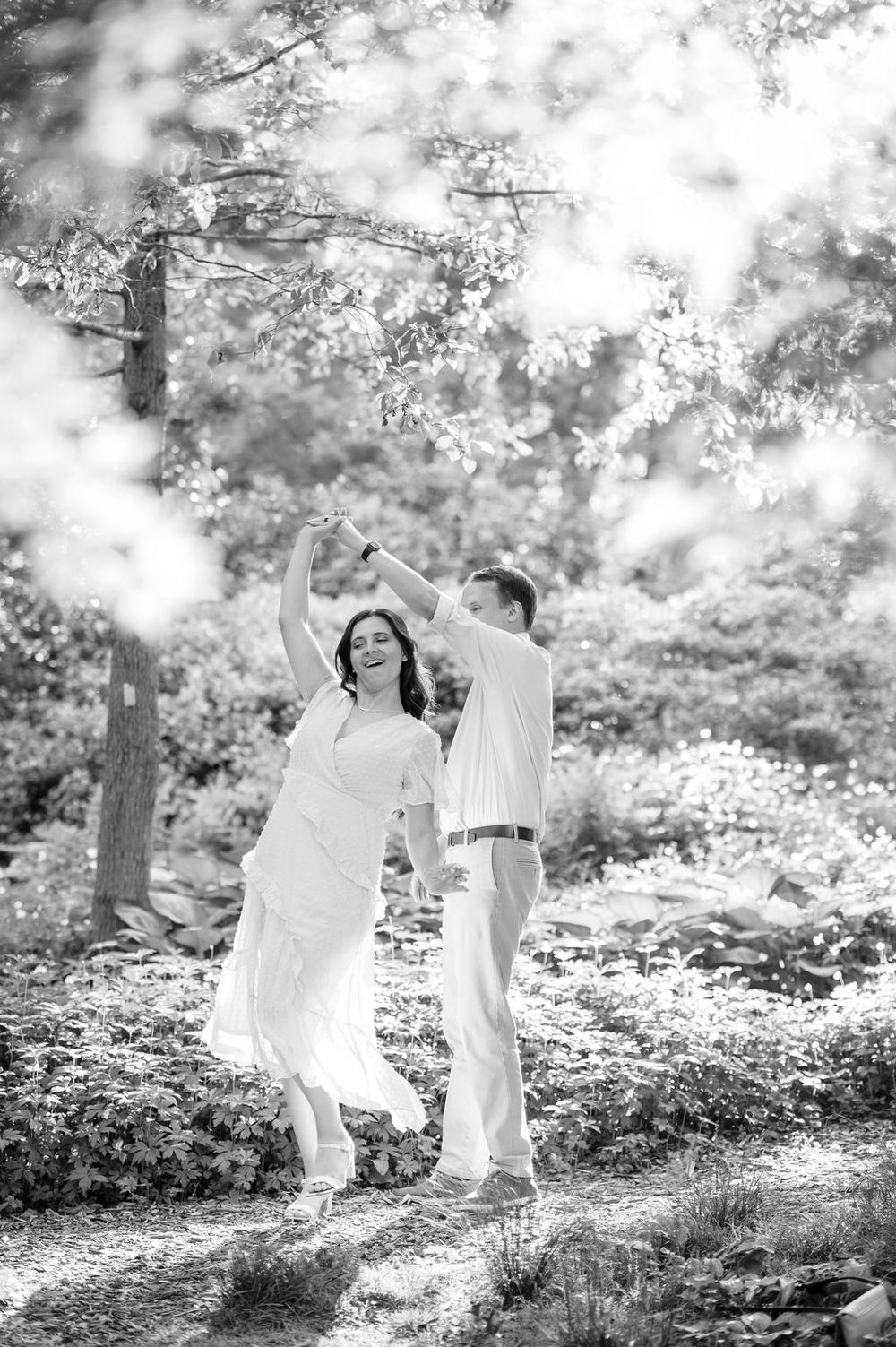 Two people in flowing white outfits dance playfully together in a dreamy black and white forest setting.