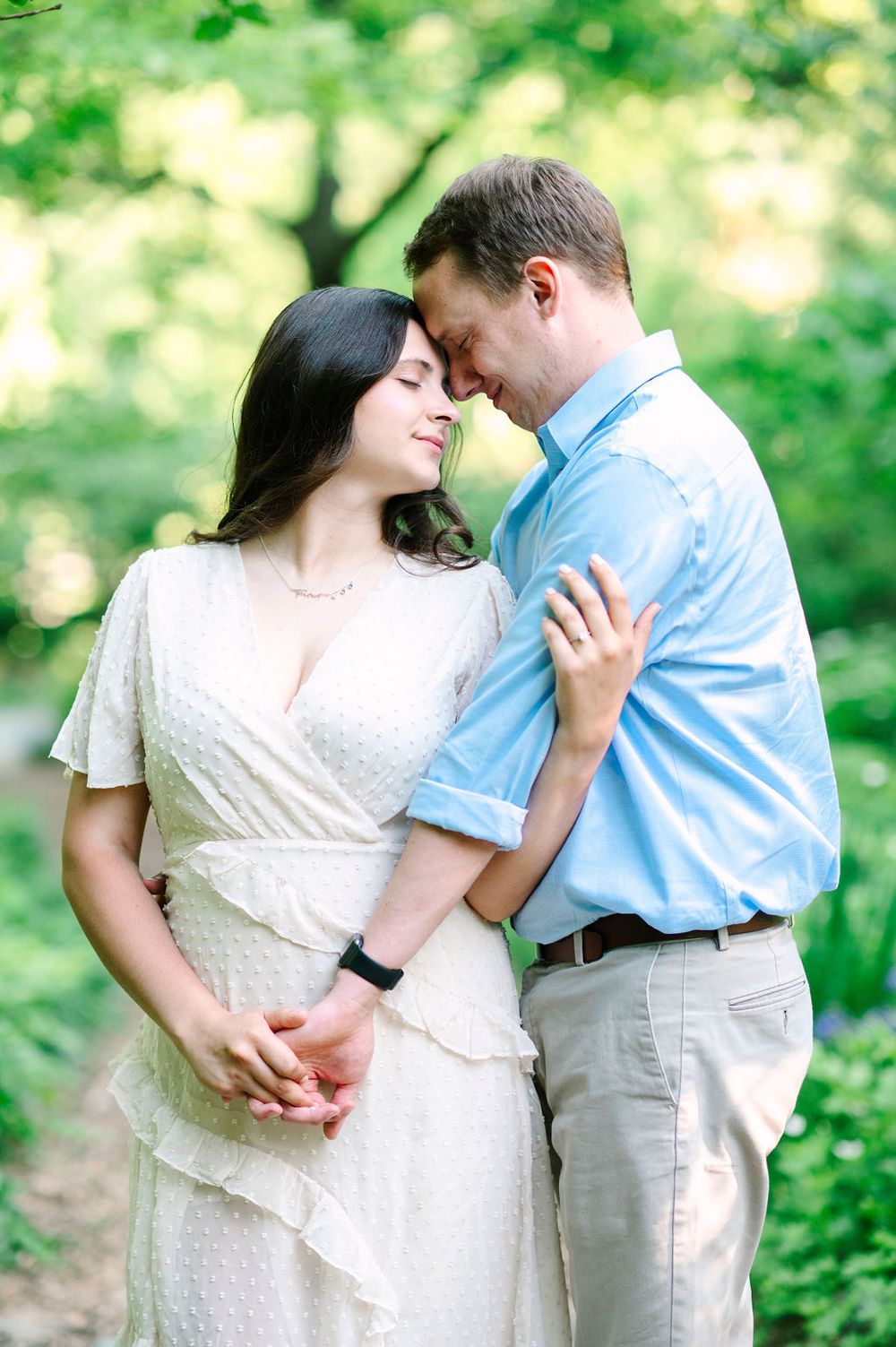 Romantic couple in white dress and blue shirt sharing intimate moment outdoors with green foliage in background.