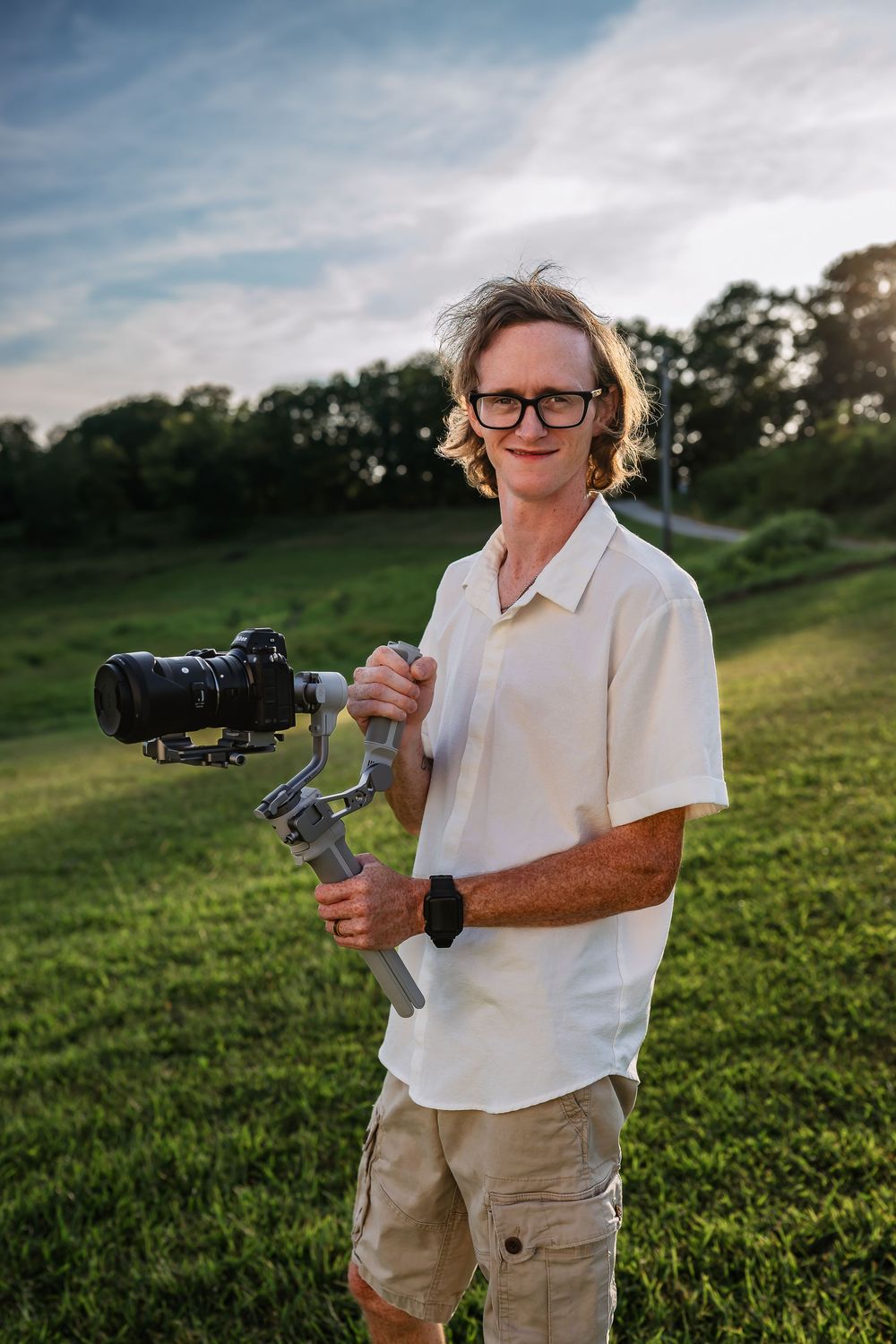 Camera operator in white shirt and khaki shorts holds professional camera rig on outdoor grassy hill at sunset.