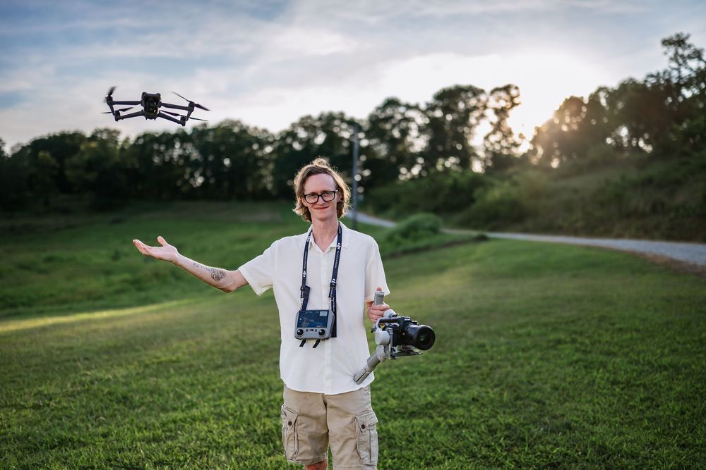 Professional drone pilot stands in a grassy field during sunset while operating camera equipment.