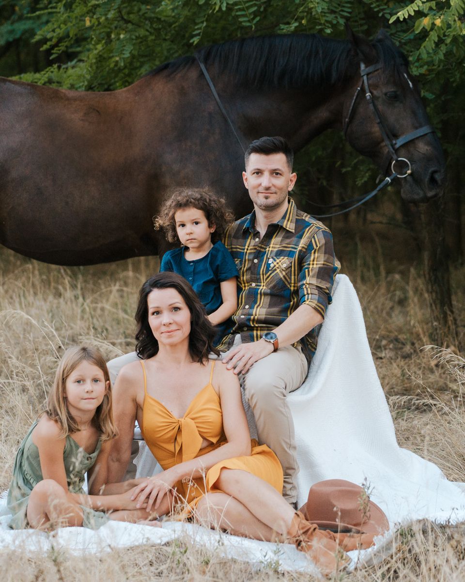 A family in neutral coloured outfits poses together on a white blanket in a outdoor setting with a horse in the background photoshoot Sydney