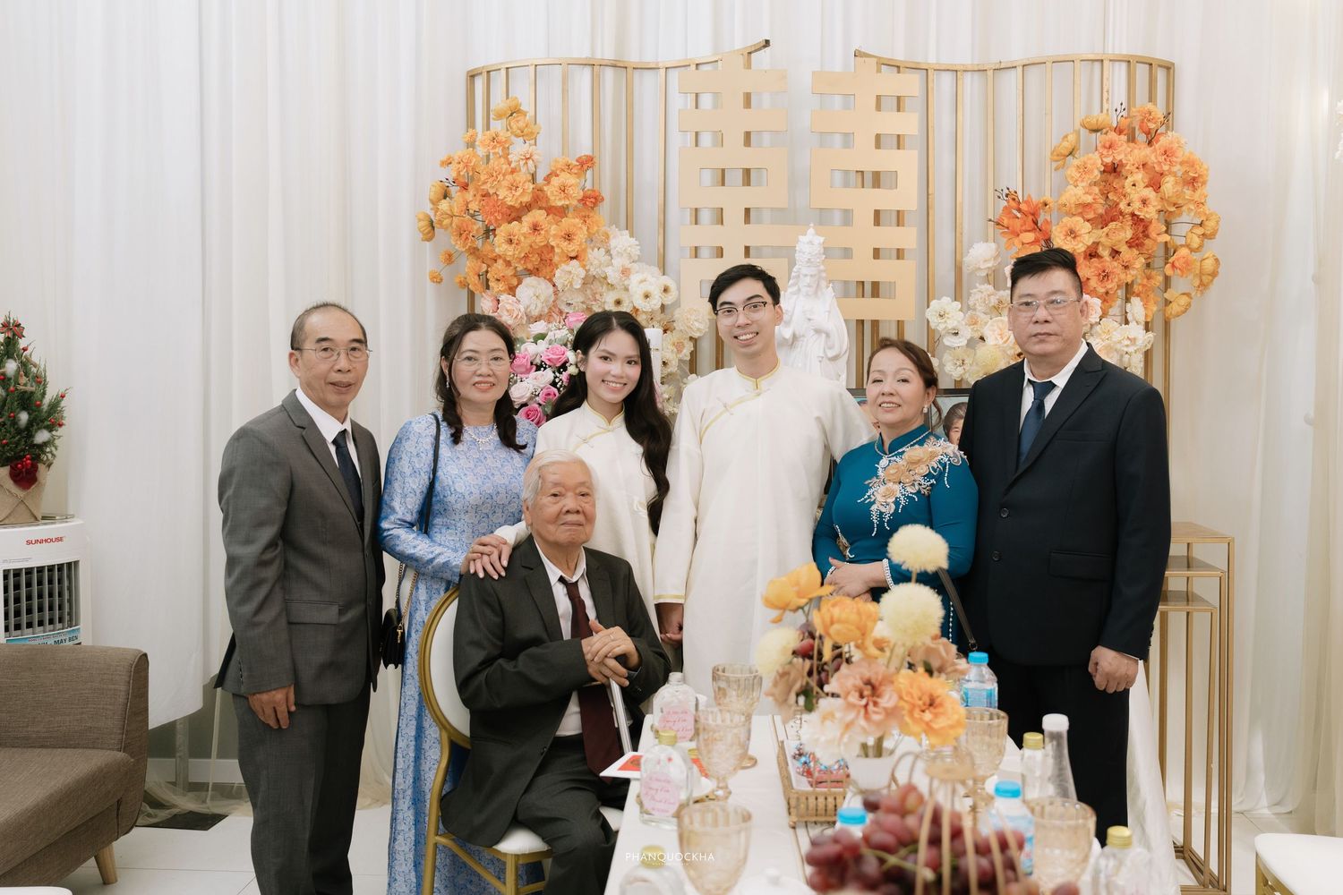Vietnamese wedding family portrait with guests in traditional ao dai attire posing before floral backdrop.