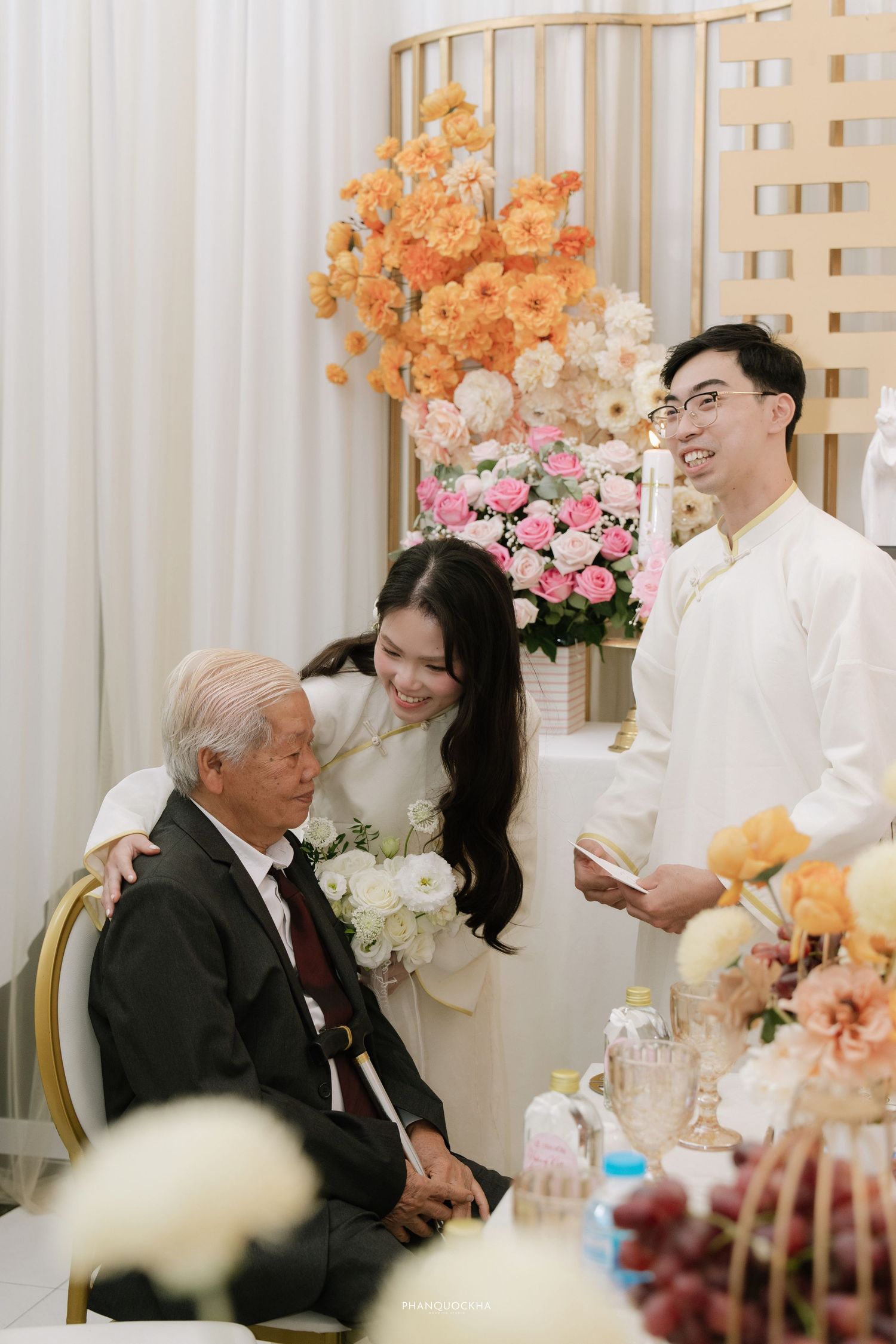 Bride and groom in white ao dai greeting elderly grandfather at Vietnamese wedding ceremony with flowers.
