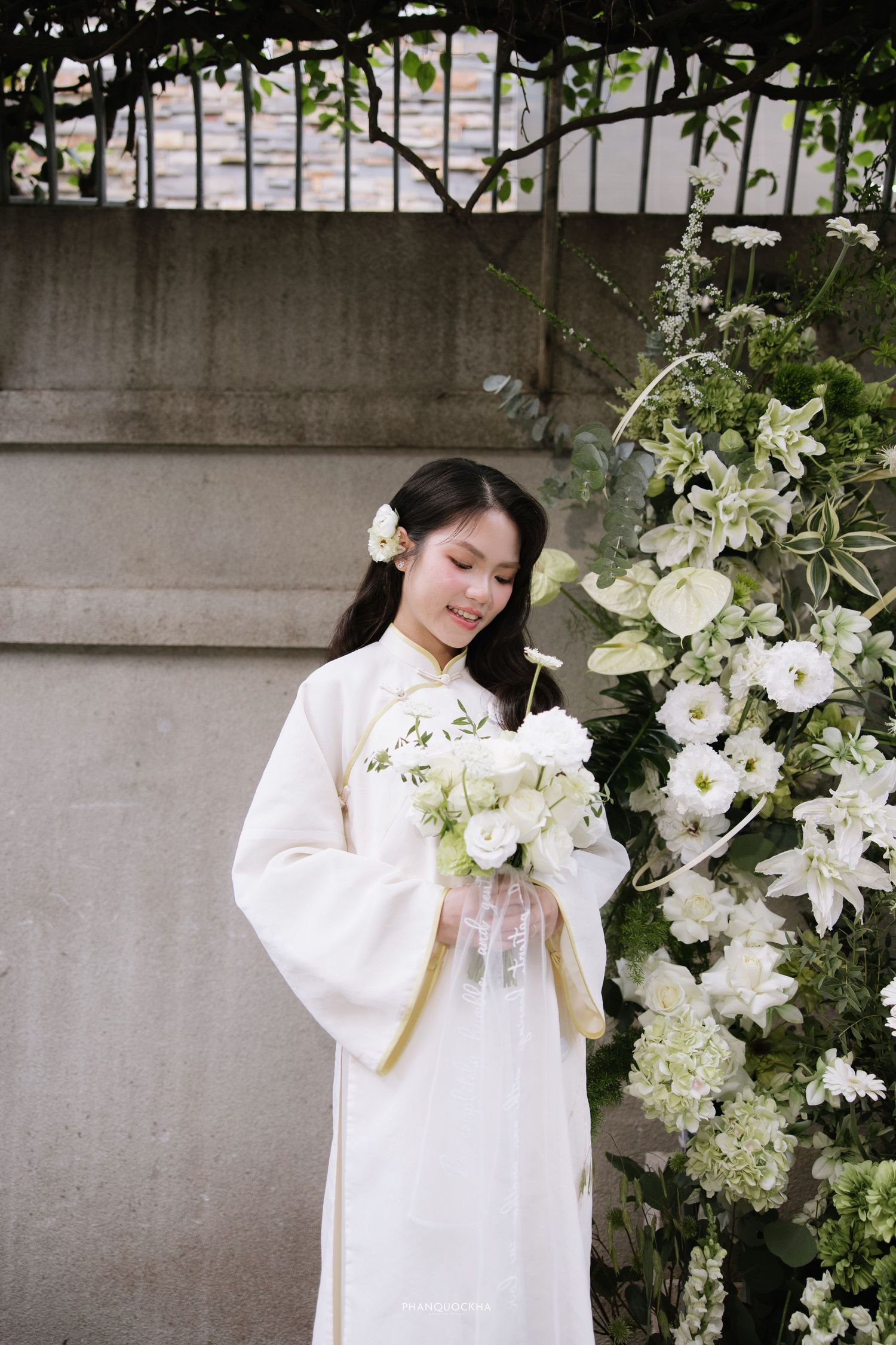 Bride in white traditional Vietnamese ao dai looking down at white flower bouquet outdoors.