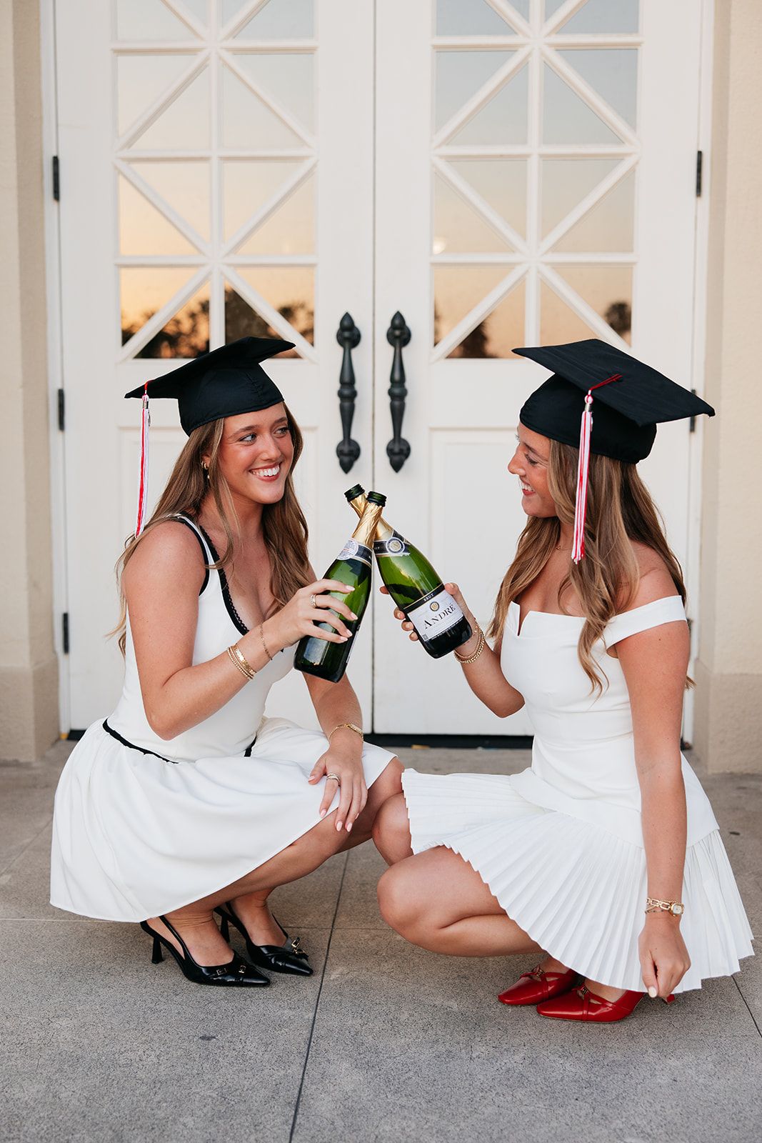Two graduates in white dresses laugh and celebrate with champagne on their graduation day.