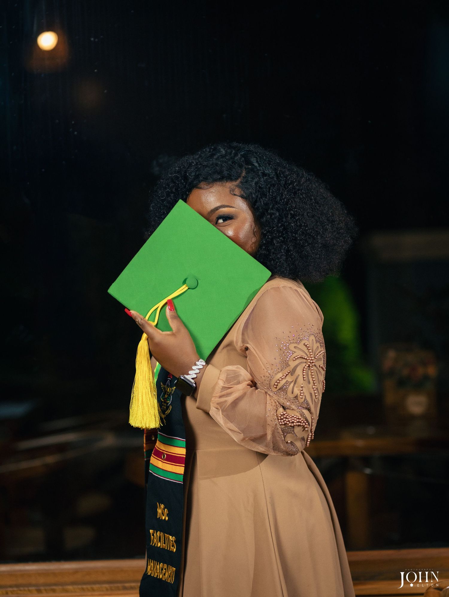 Graduate student in cap and gown poses confidently against modern interior railing with ambient lighting.