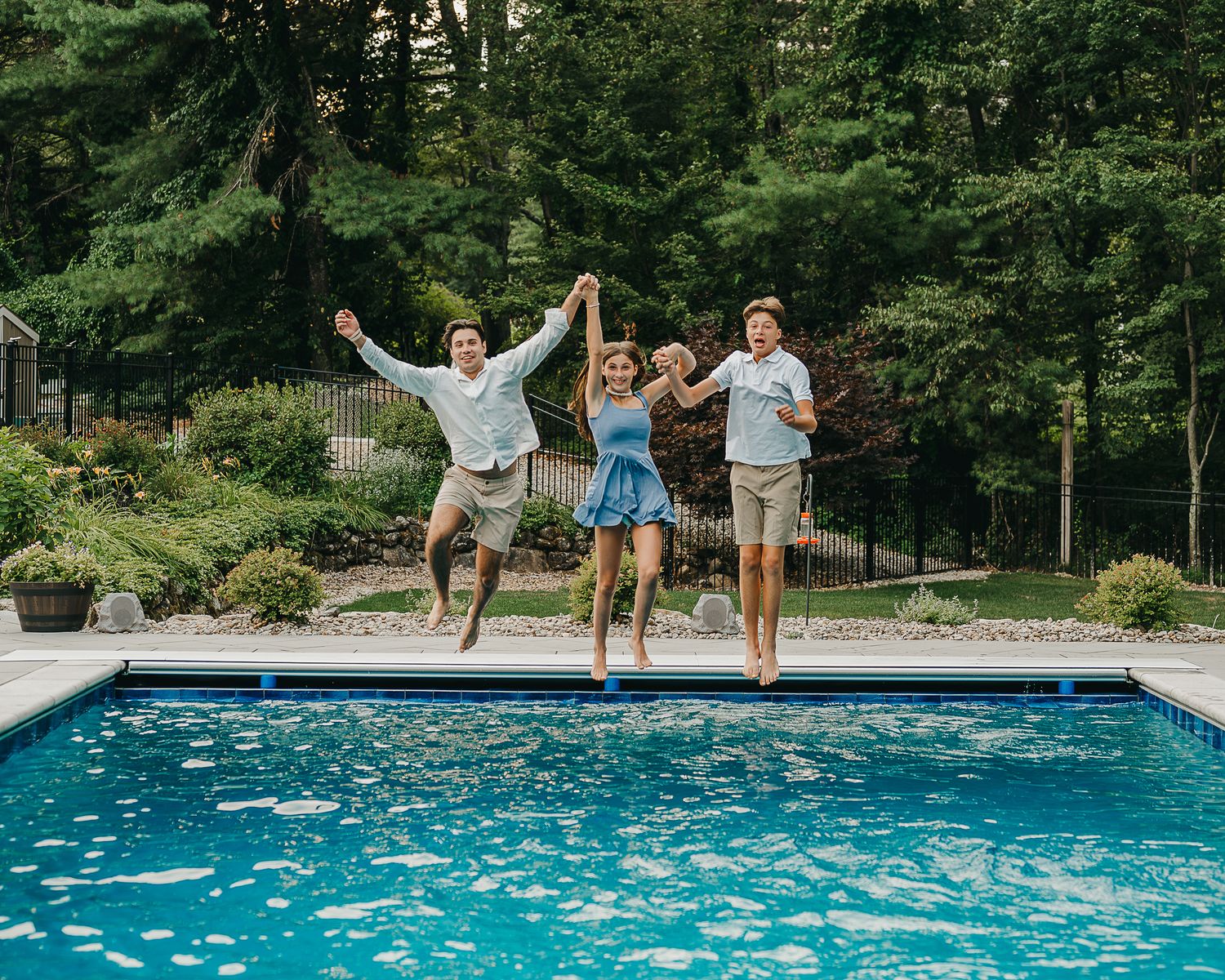Three teenage siblings jump in to a pool together during a portrait session in summer with trees in the background.
