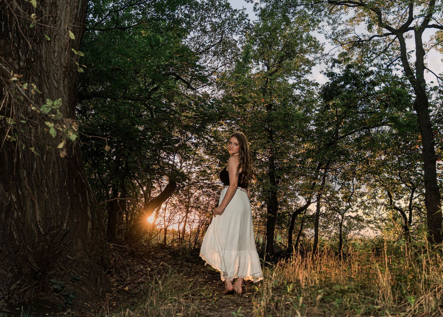 Woman in flowing white dress stands in forest path during golden sunset through trees.