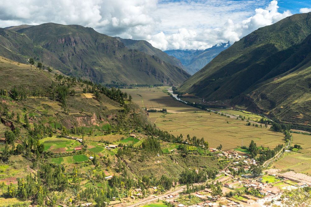 Sweeping mountain valley landscape showcases green fields and winding roads beneath dramatic clouds in Peru's Sacred Valley.