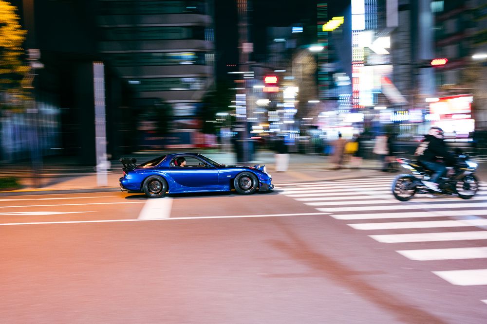 Motion-blurred blue sports car crossing an illuminated intersection in Tokyo at night.