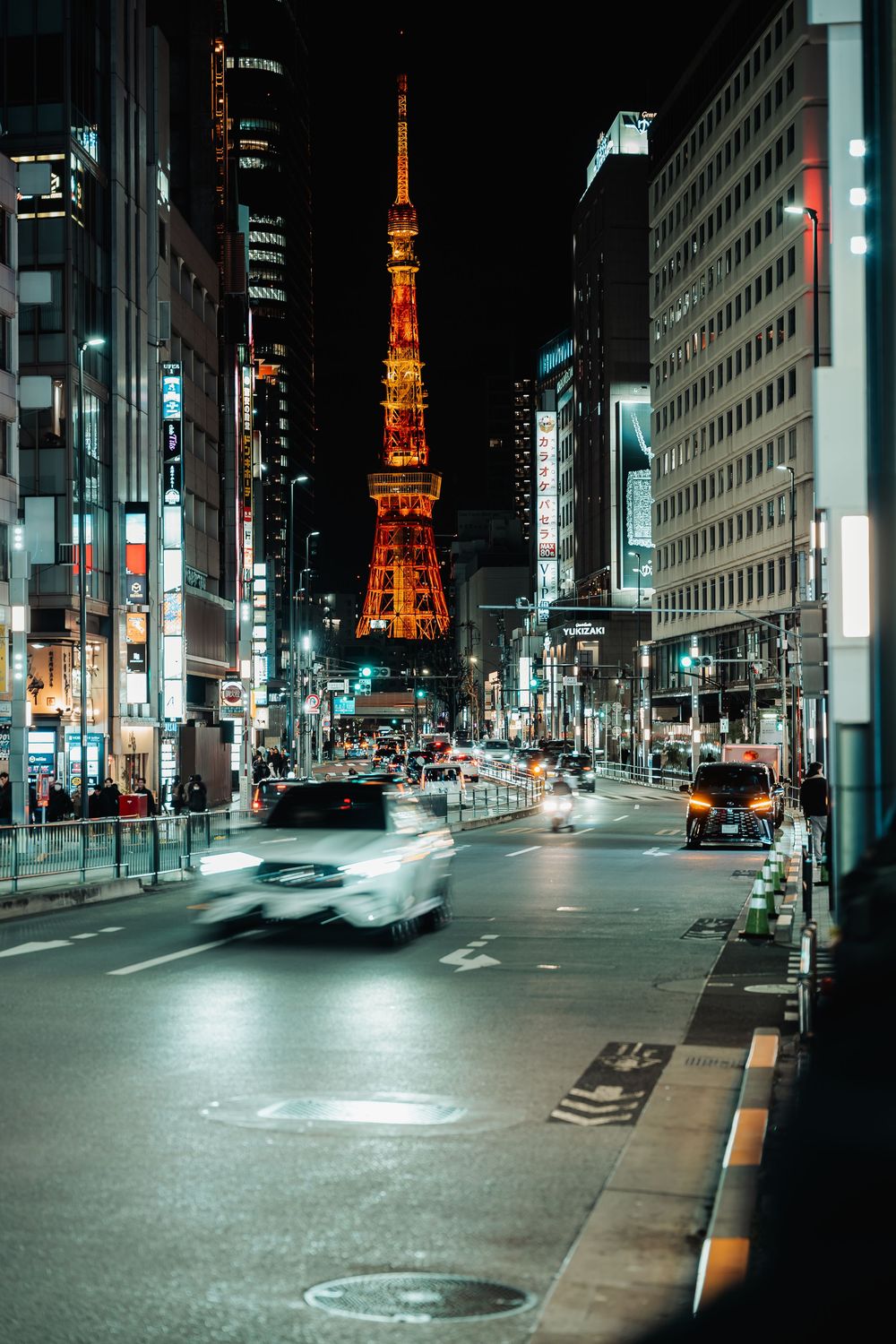 Illuminated Tokyo Tower glows orange against the night sky as seen from an empty city street with modern buildings.
