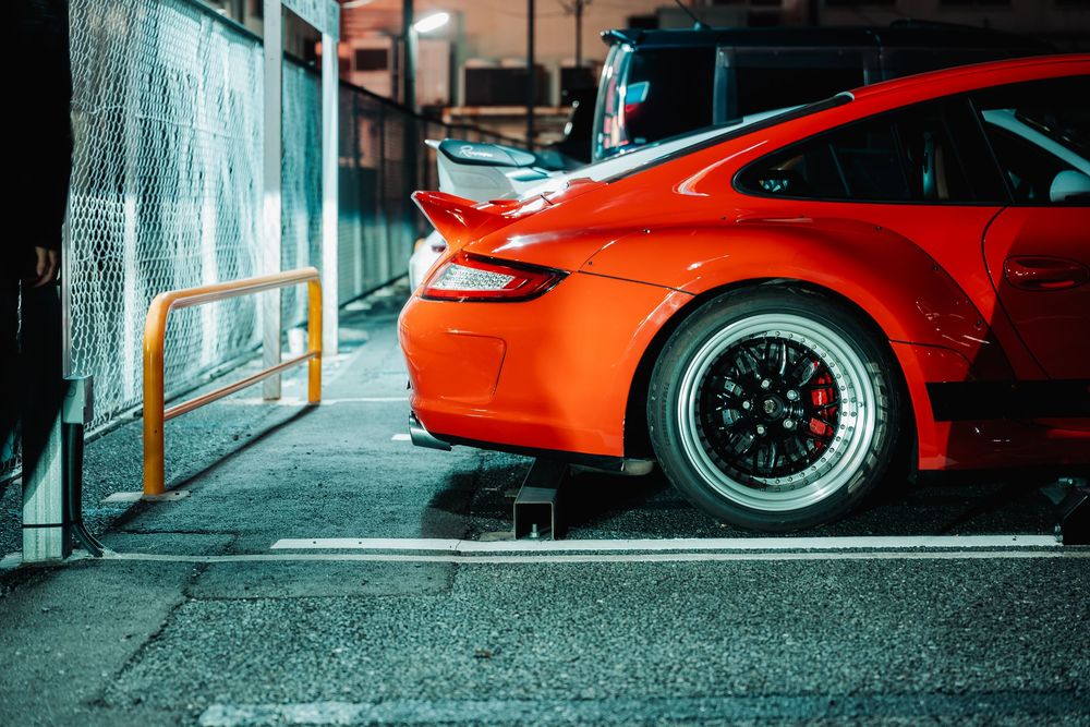 Close-up view of a bright orange Porsche 911 with wide body kit and black BBS wheels parked in a garage.