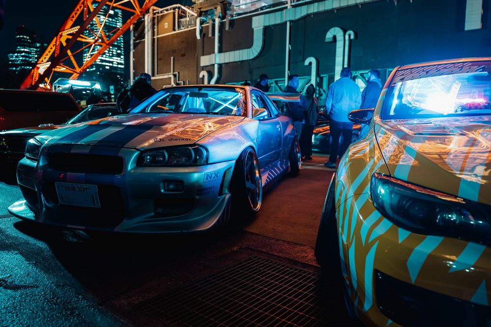 Modified Nissan Skyline GT-R and police car illuminated by neon lights at night under a highway overpass.