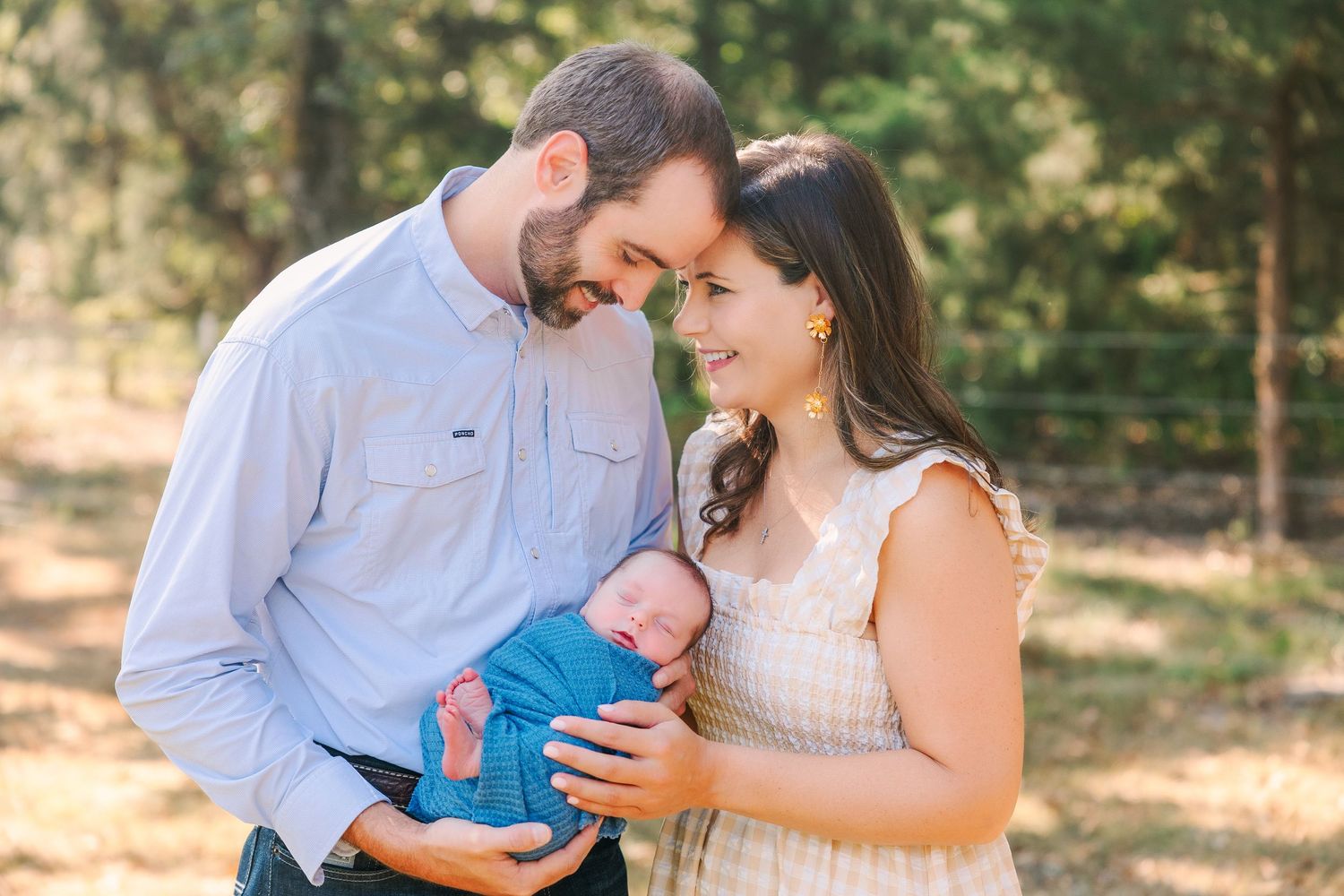 A couple in matching light blue and cream outfits share tender moments together with their baby outdoors in nature.