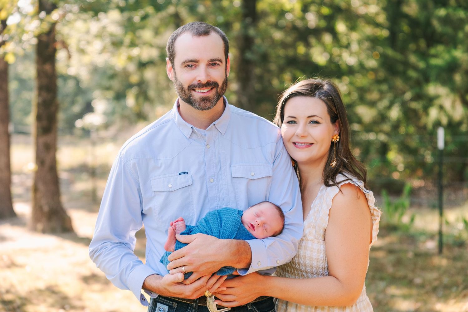 A family stands together outdoors in a sunlit wooded area while holding their newborn baby wrapped in blue.