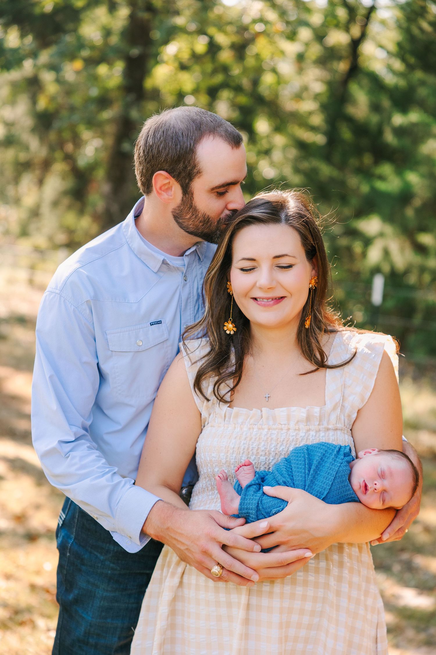 Family poses together outdoors for candid portraits in a sunny wooded setting wearing casual clothes.