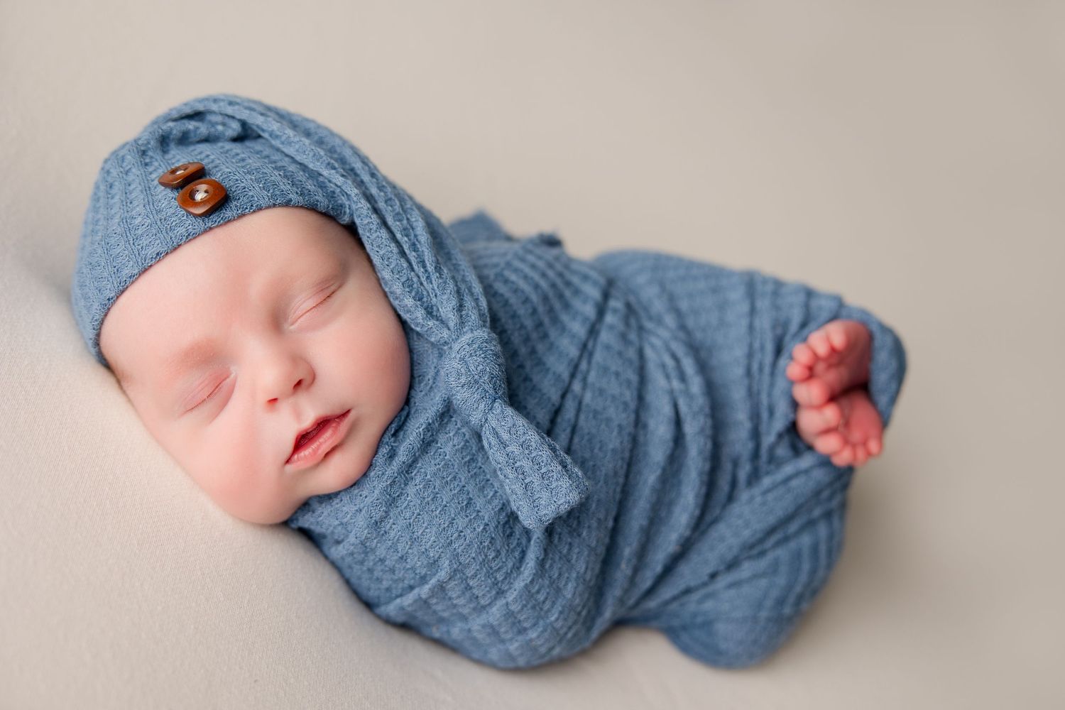 Sleeping newborn baby wrapped in soft blue swaddle blanket lying on beige background.