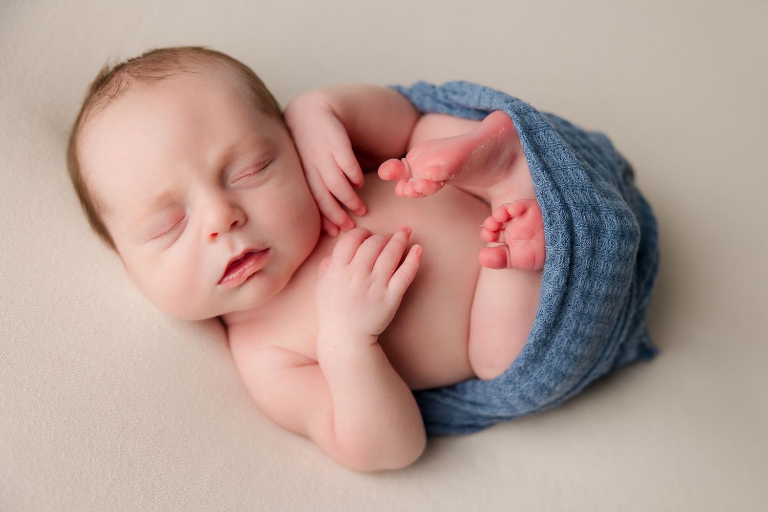 Sleeping baby wearing blue knit hat lies peacefully on soft cream-colored background in newborn photography session.